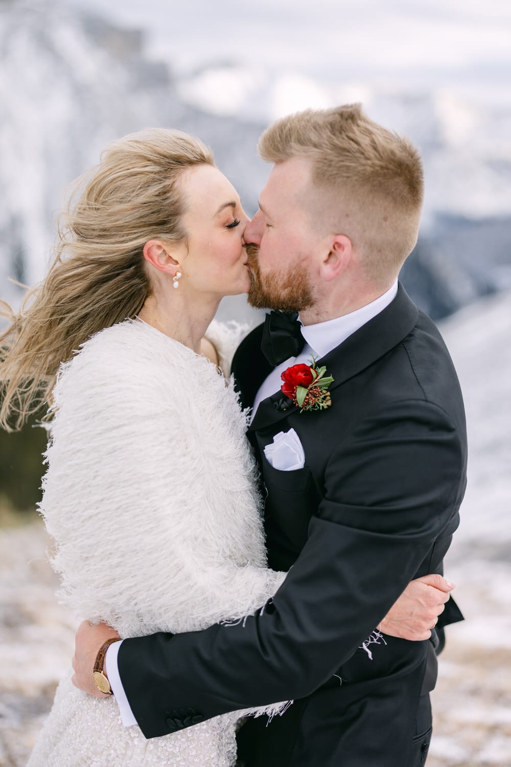 A bride and groom sharing a kiss in a snowy mountain setting, with the bride wearing a white fur shawl and the groom in a black suit with a red boutonniere.