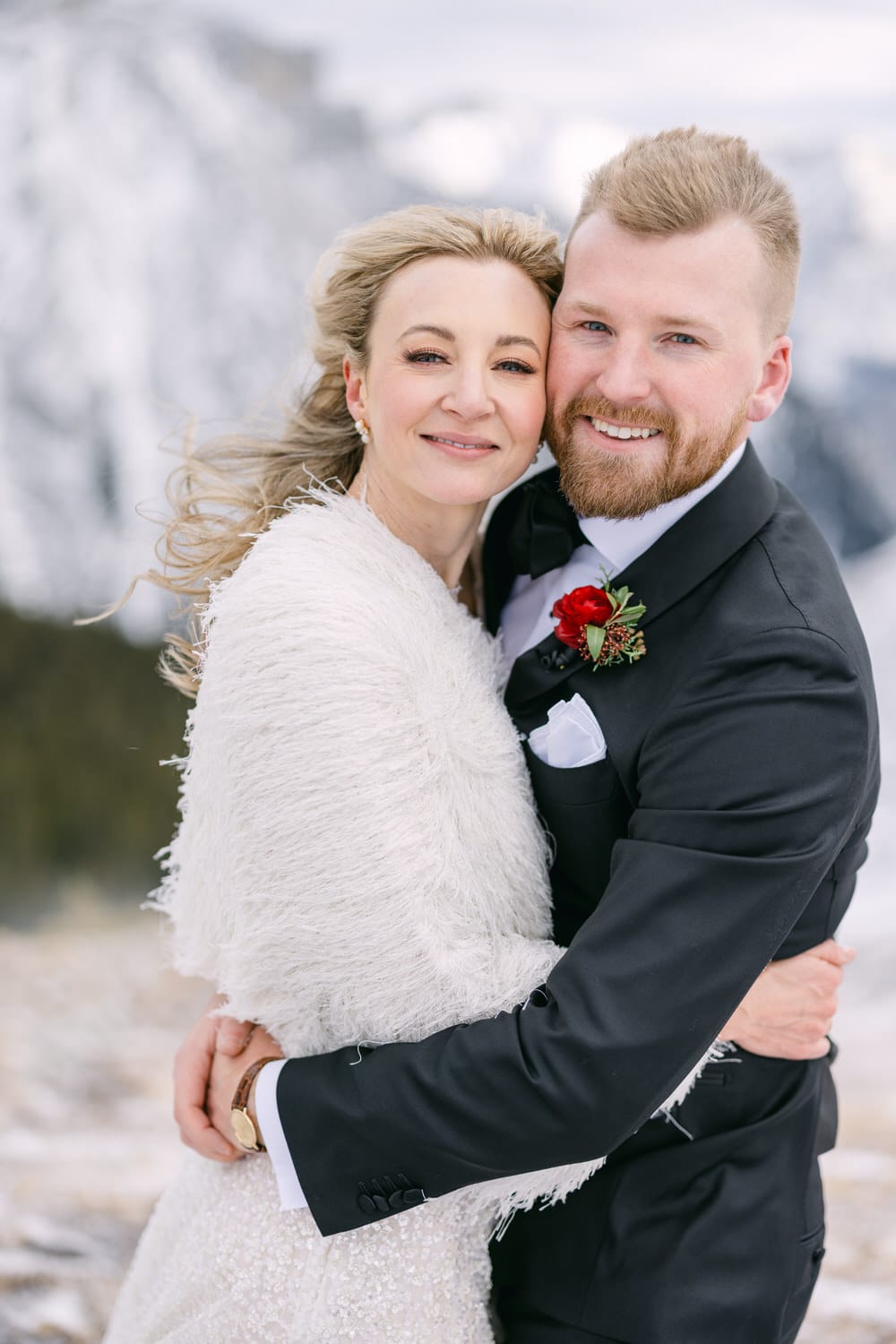 A smiling couple in wedding attire hugging against a snowy mountain backdrop.