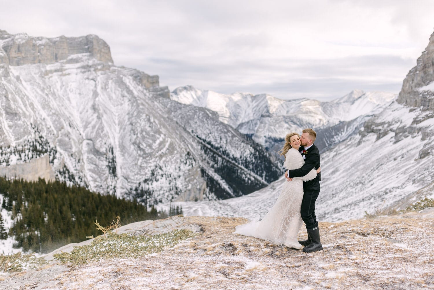 A couple in wedding attire sharing a kiss on a mountain summit with snow-covered peaks in the background