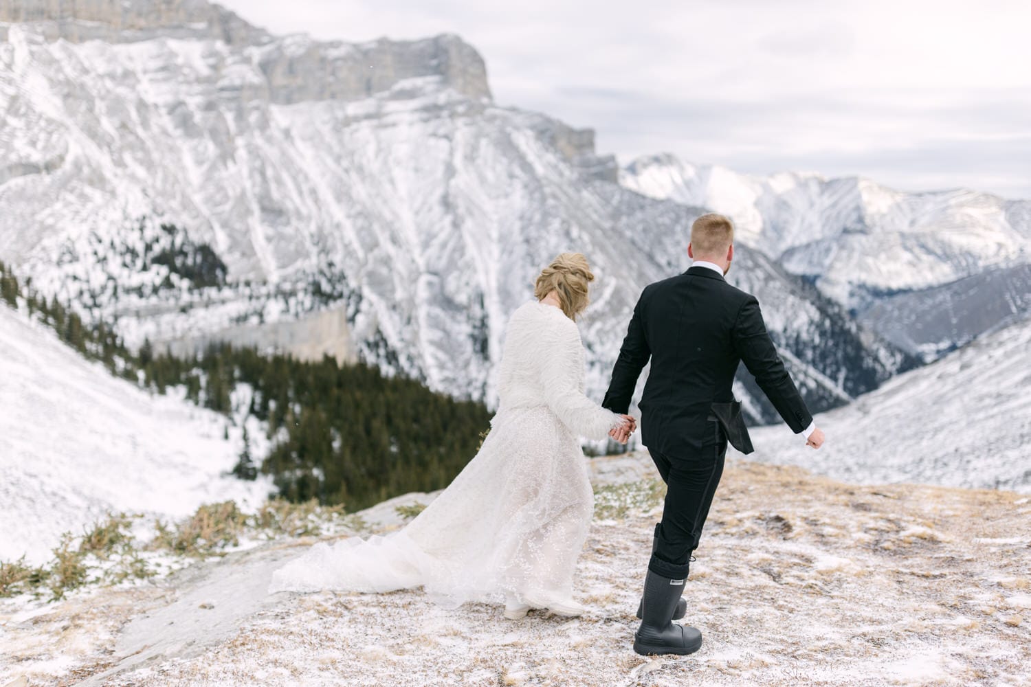 A couple in wedding attire holding hands against a backdrop of snowy mountains, with the bride in a white dress and the groom in a black suit.