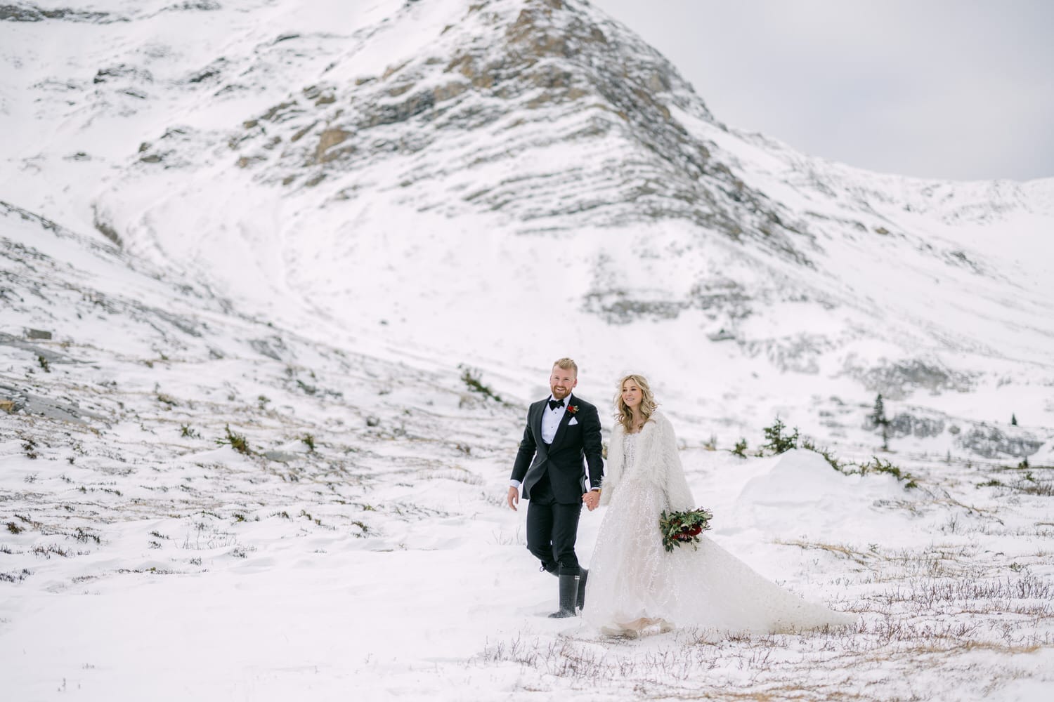 A couple in wedding attire holding hands amidst a snowy mountain landscape.