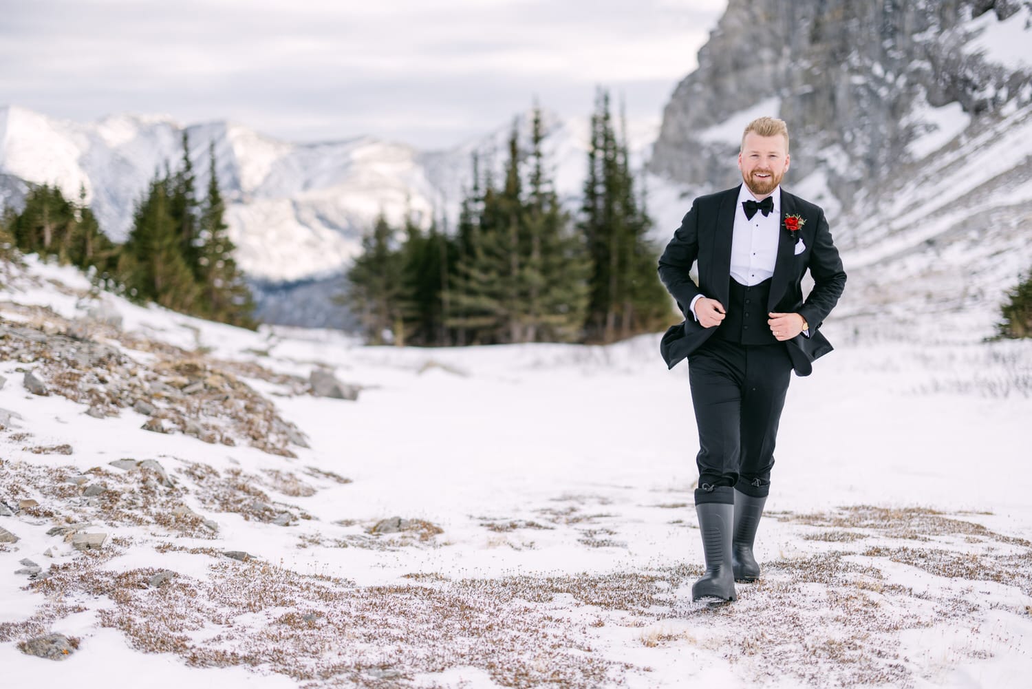 Man in a tuxedo with a boutonniere walking in a snowy mountain landscape.