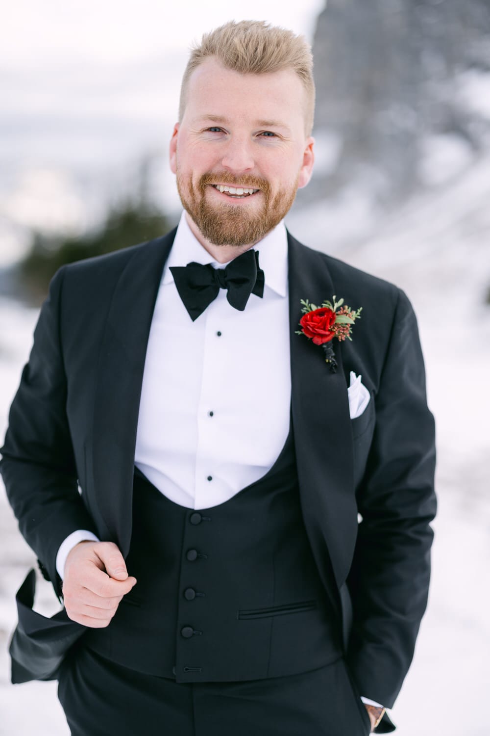 A smiling man with a beard wearing a black tuxedo with a bow tie and a red rose boutonnière, standing against a snowy backdrop.