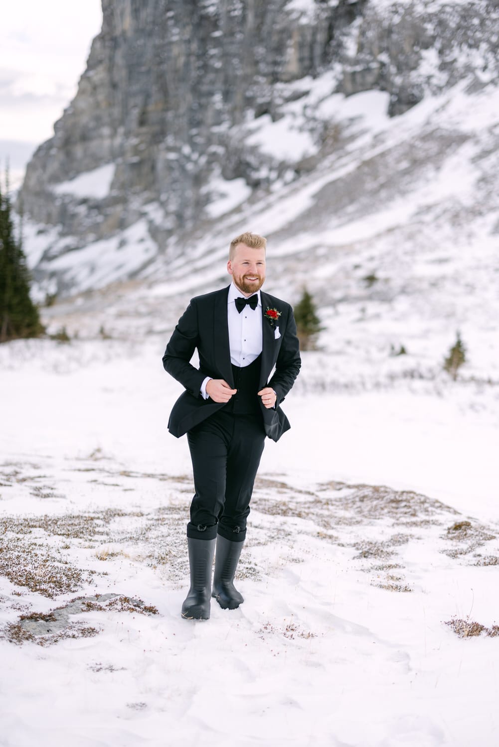 A man in a tuxedo with a bow tie and rubber boots standing in the snow with a mountainous backdrop.