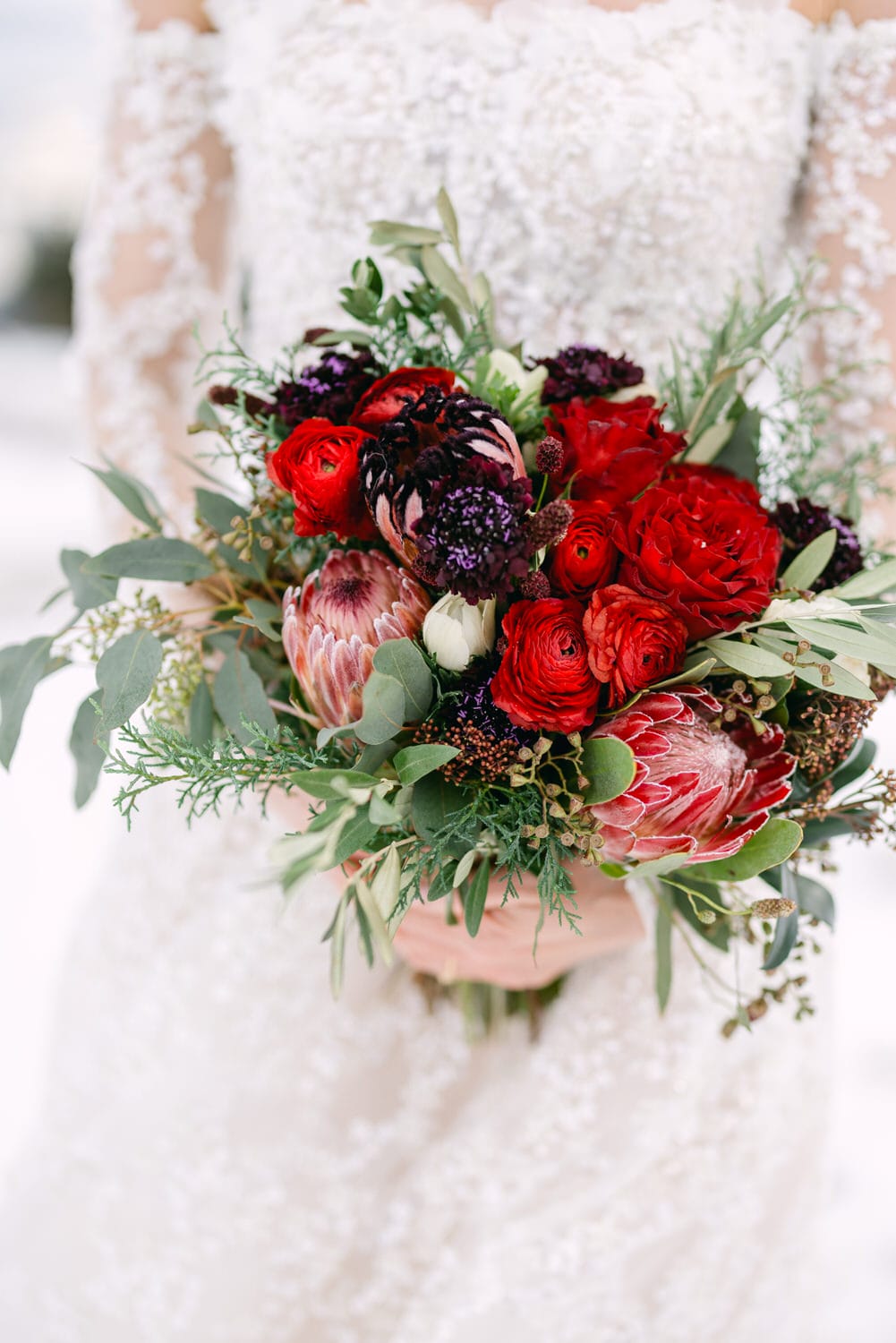 A close-up of a bride holding a colorful bouquet with red roses, dark flowers, and green foliage, complemented by the intricate details of her white beaded wedding dress.