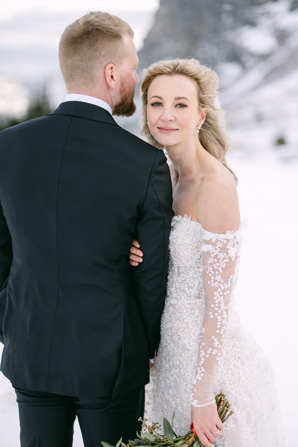 Bride in a lace dress smiling at the camera while embracing groom in a black suit against a snowy backdrop