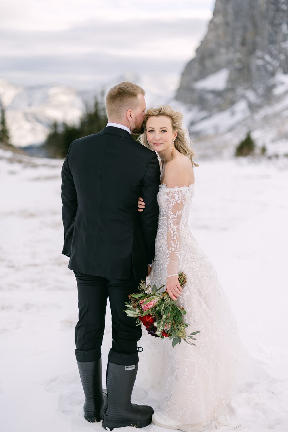 A bride and groom embracing in a snowy landscape, with the bride in a white lace dress holding a bouquet and the groom in a black suit with rubber boots.