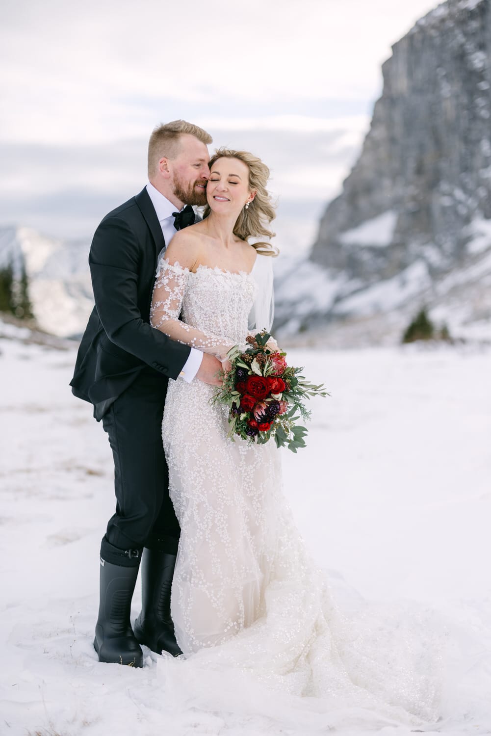 A bride and groom sharing a loving embrace in a snowy landscape with mountain backdrop.