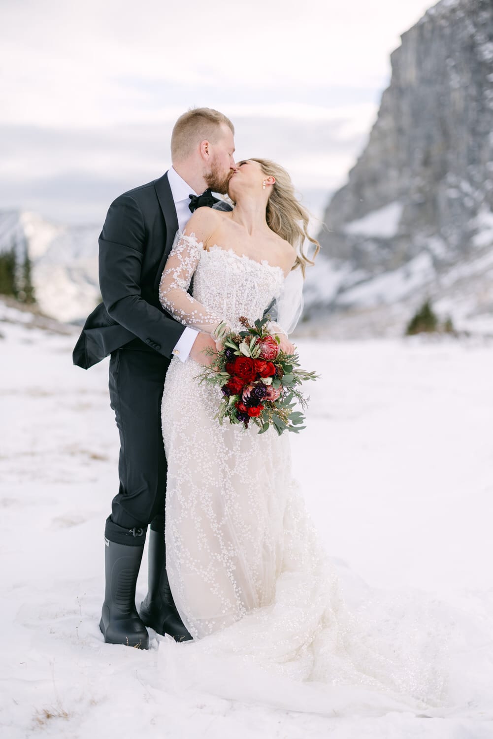 A bride and groom kissing in a snowy landscape with mountains in the background.