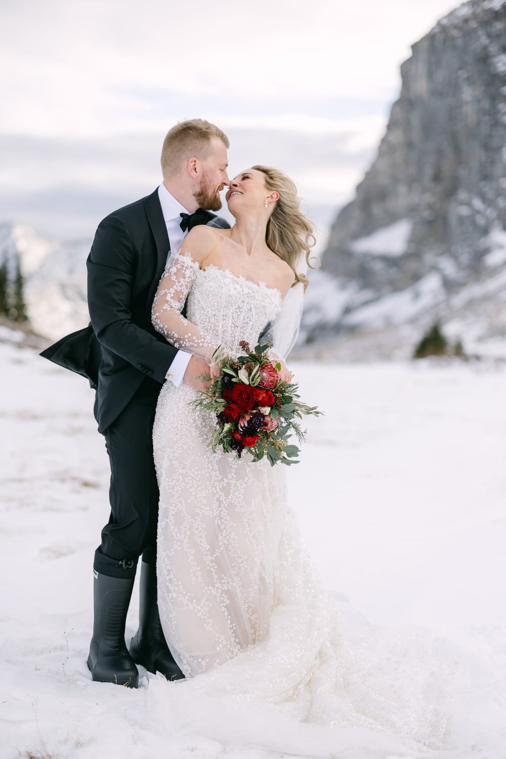 A couple in wedding attire sharing a kiss in a snowy landscape with mountains in the background.