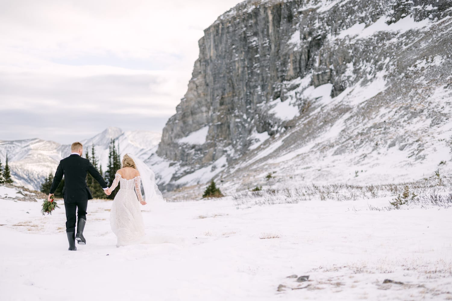 A bride and groom holding hands and walking through snow with a cliff and mountains in the background.