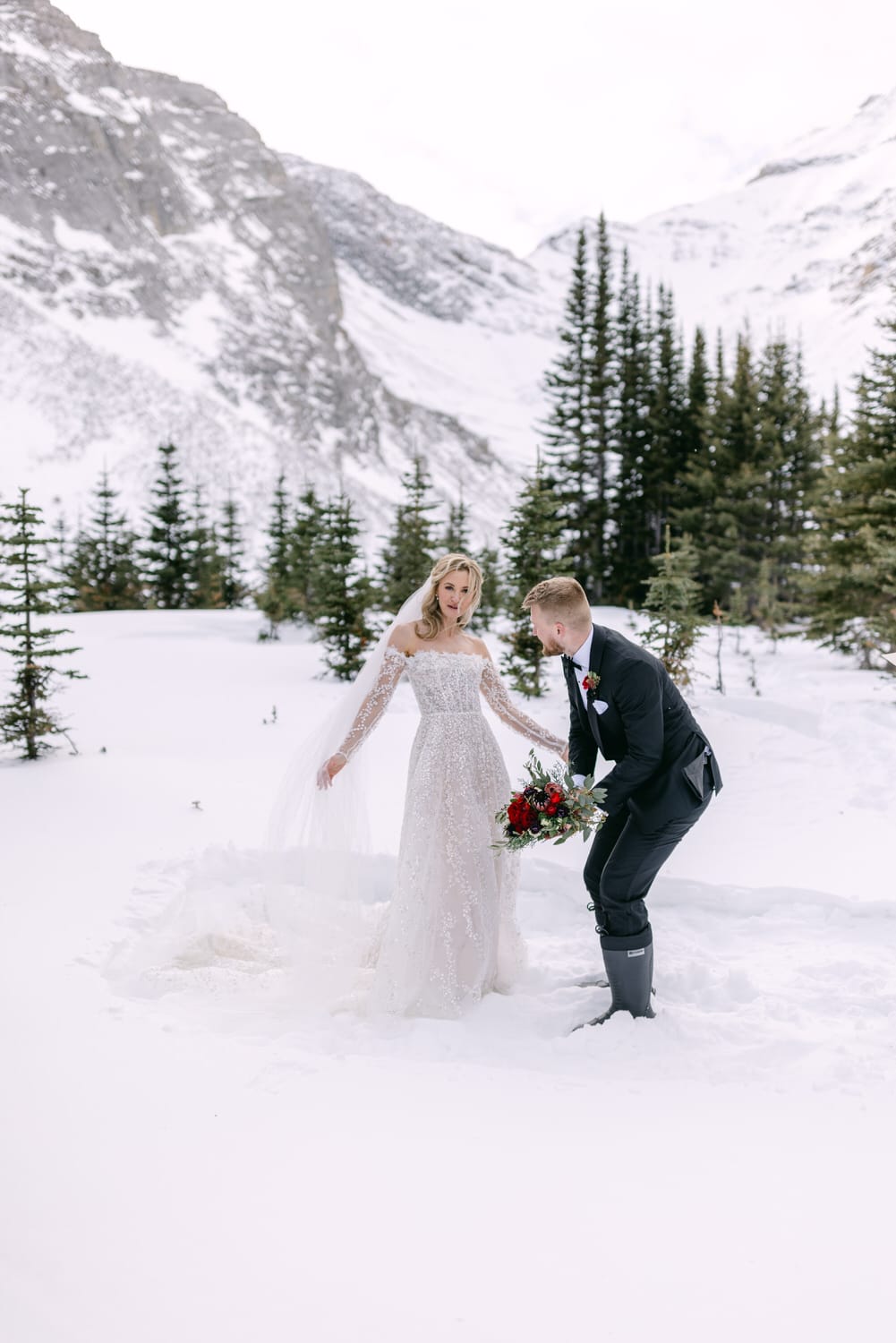 A bride in a white lace dress and a groom in a black suit sharing a playful moment in a snowy mountain landscape.