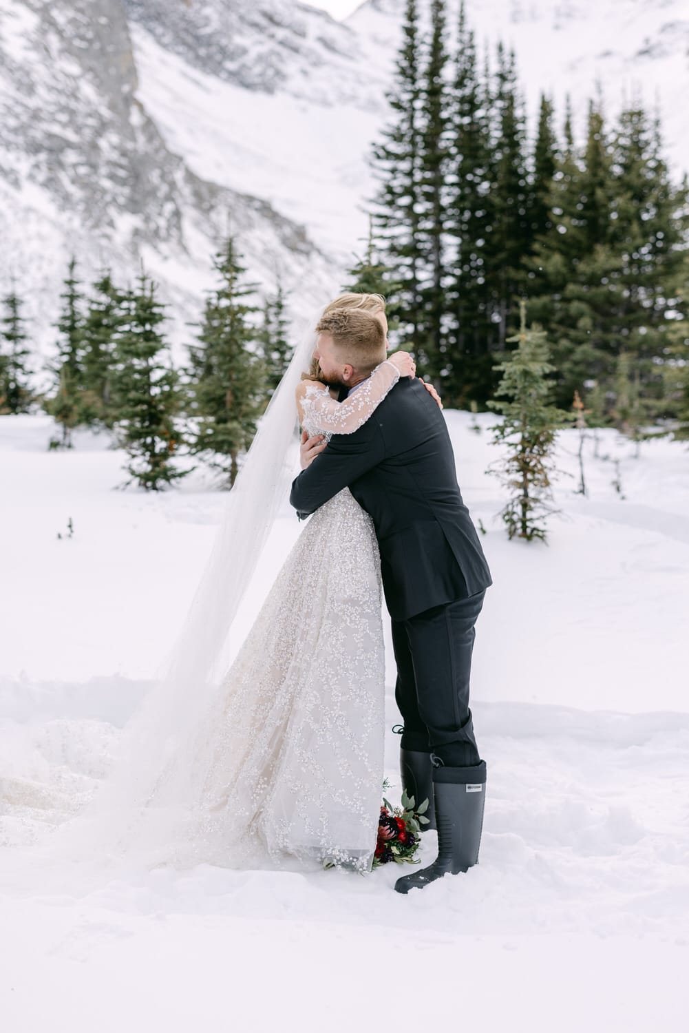 A bride and groom hugging each other in a snow-covered landscape, with pine trees and mountains in the background.