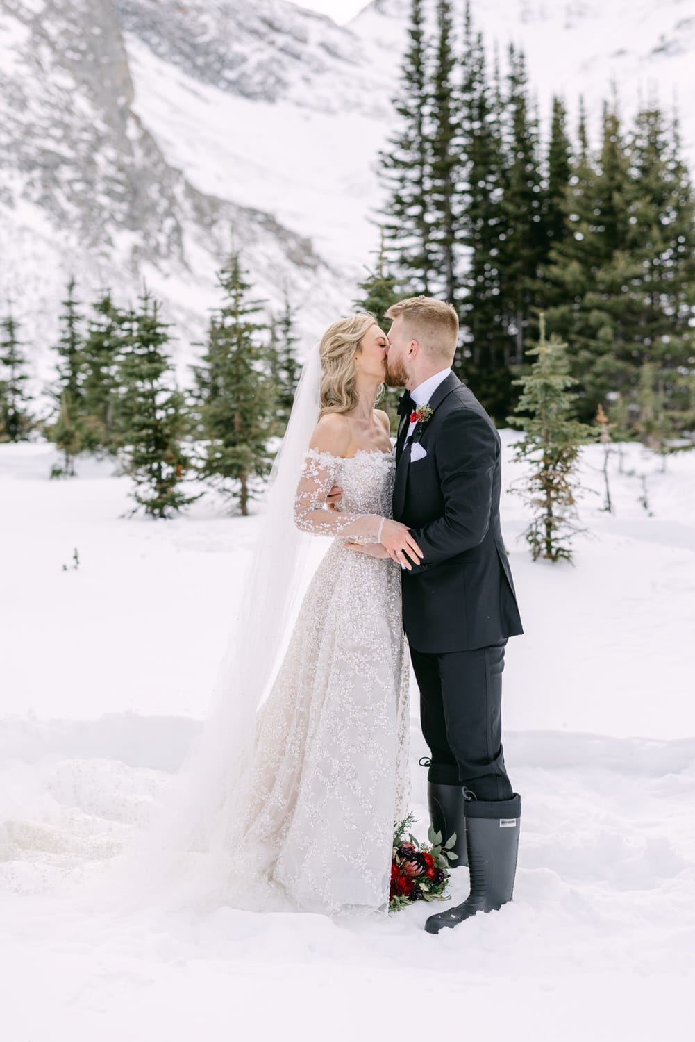 A bride and groom sharing a kiss in a snowy mountain landscape, surrounded by evergreen trees.