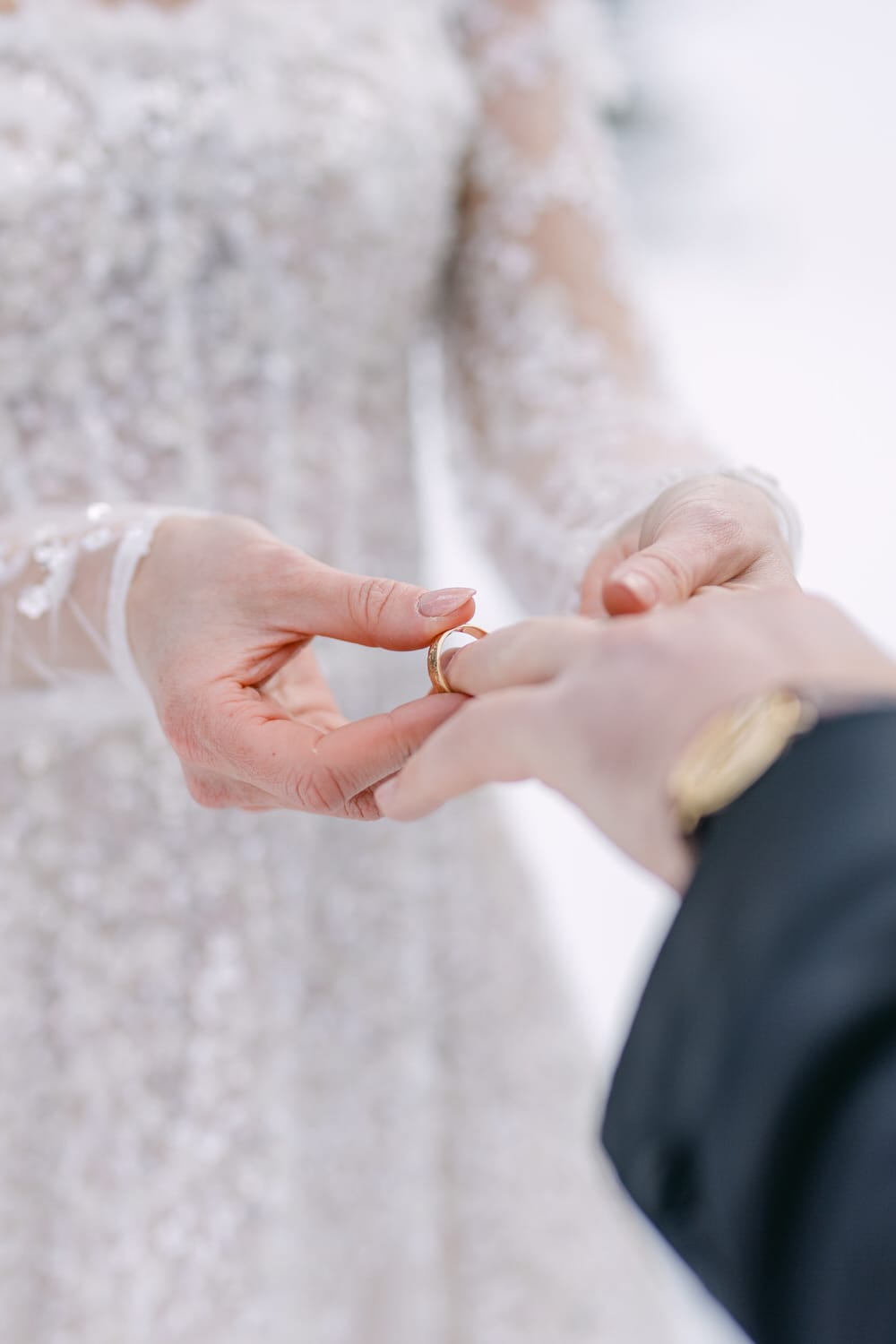 Close-up of a couple's hands as one slides a wedding ring onto the other's finger, with focus on the hands and the ring against the backdrop of a white bridal dress.