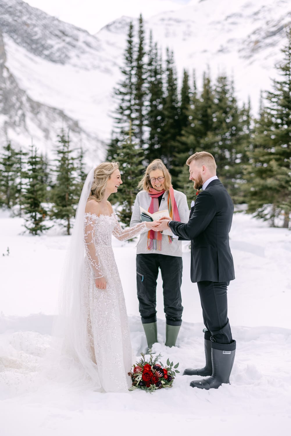 A bride and groom exchanging vows in the snow with an officiant, surrounded by pine trees and mountains.