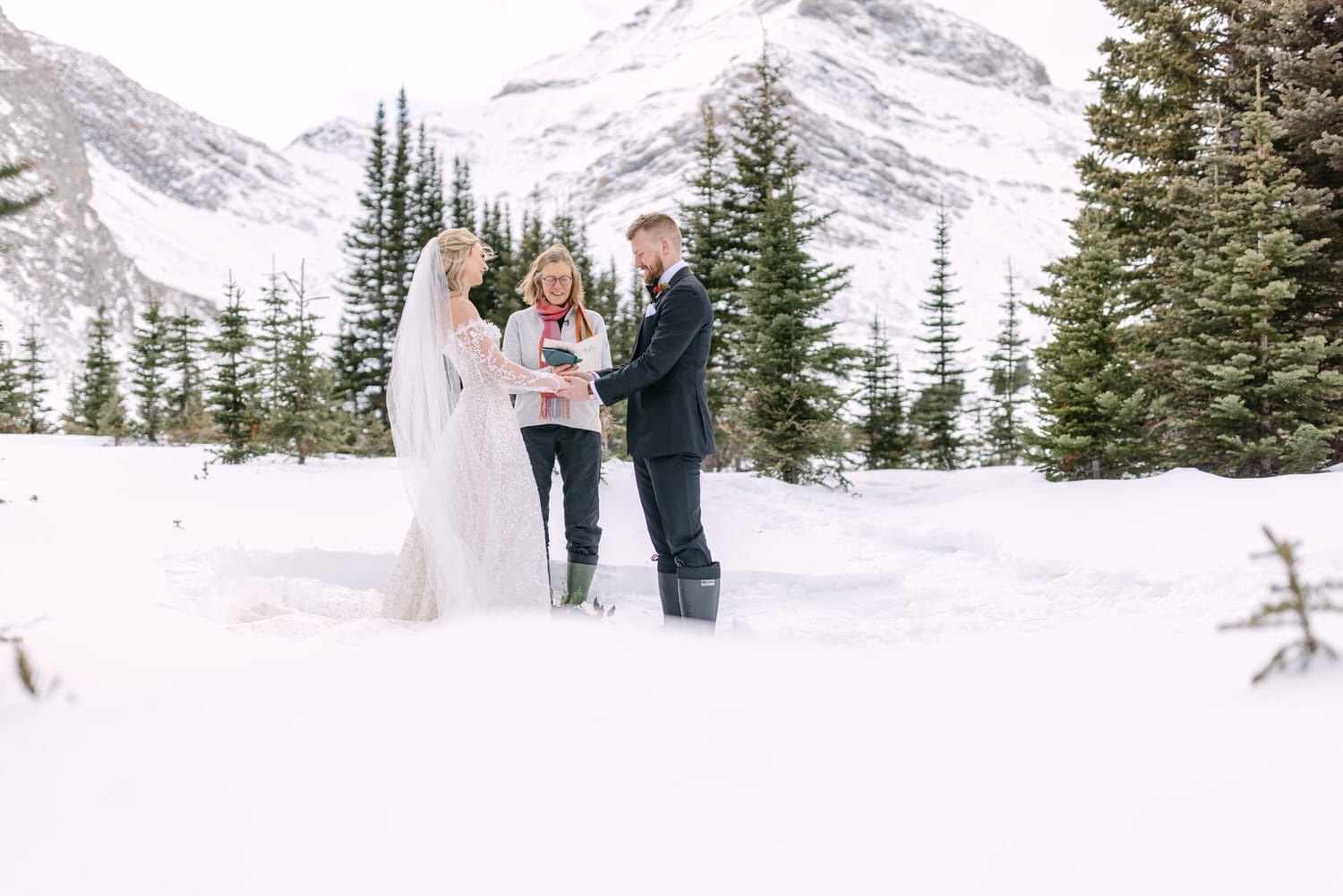 A bride and groom exchanging vows outdoors with an officiant in a snowy mountain landscape, surrounded by evergreen trees.