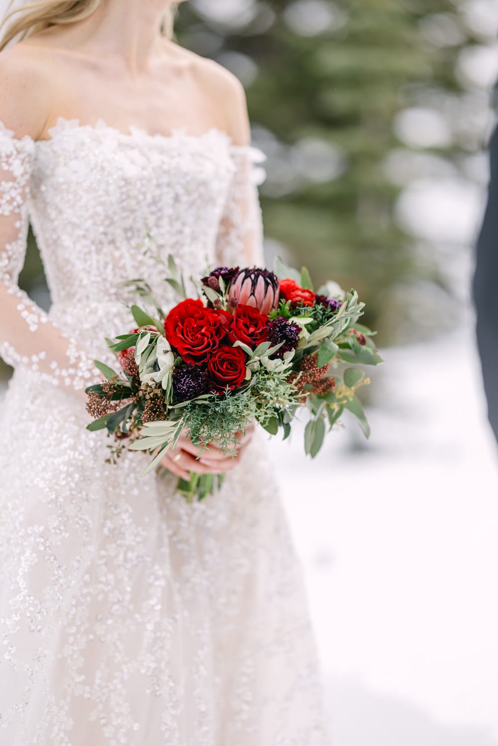 Close-up of a bride in a lace wedding dress holding a bouquet with red roses and dark botanicals against a snowy backdrop.