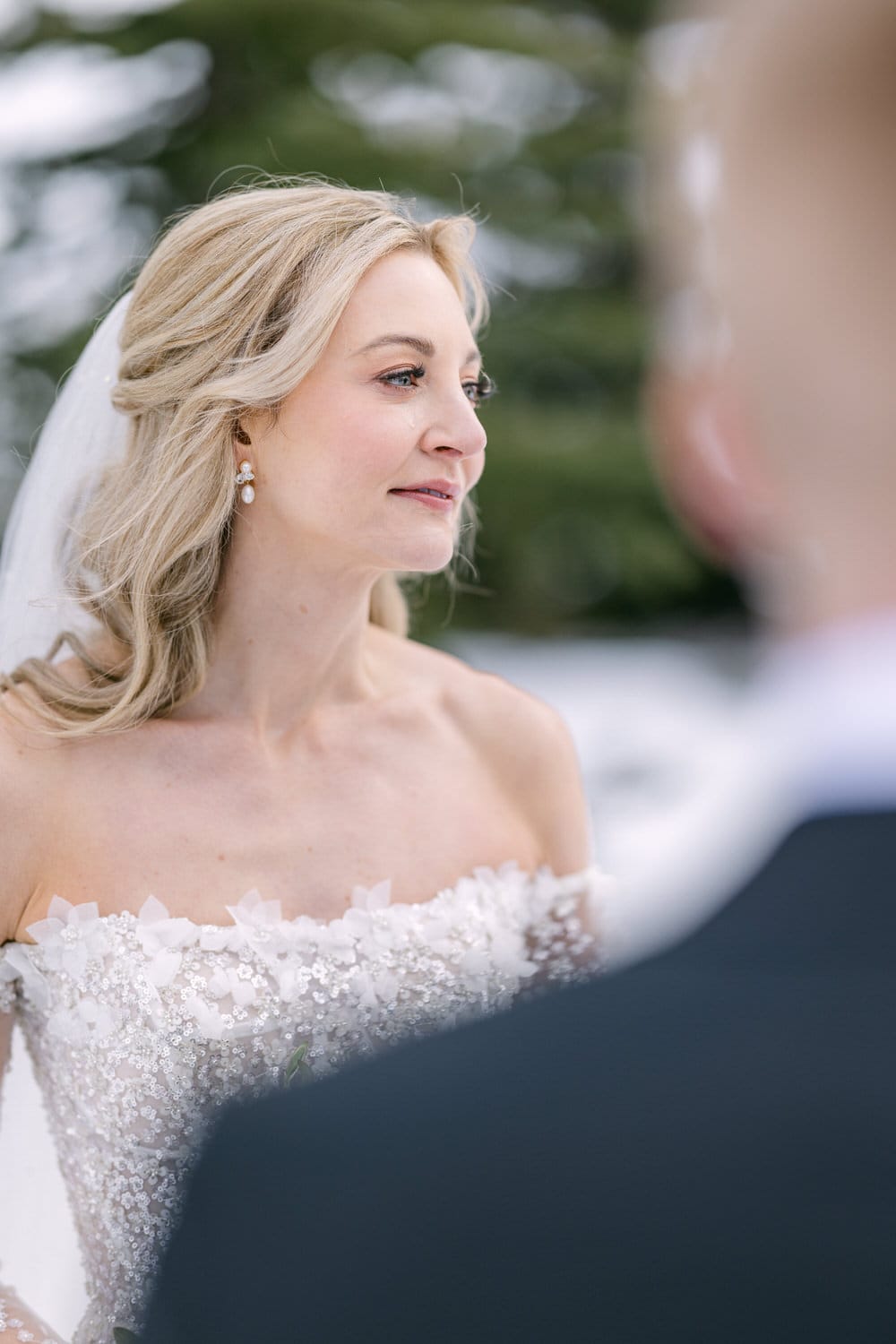 A bride in a detailed wedding gown with a smile gazing lovingly at her groom, with a blurred background indicating a wedding ceremony setting.