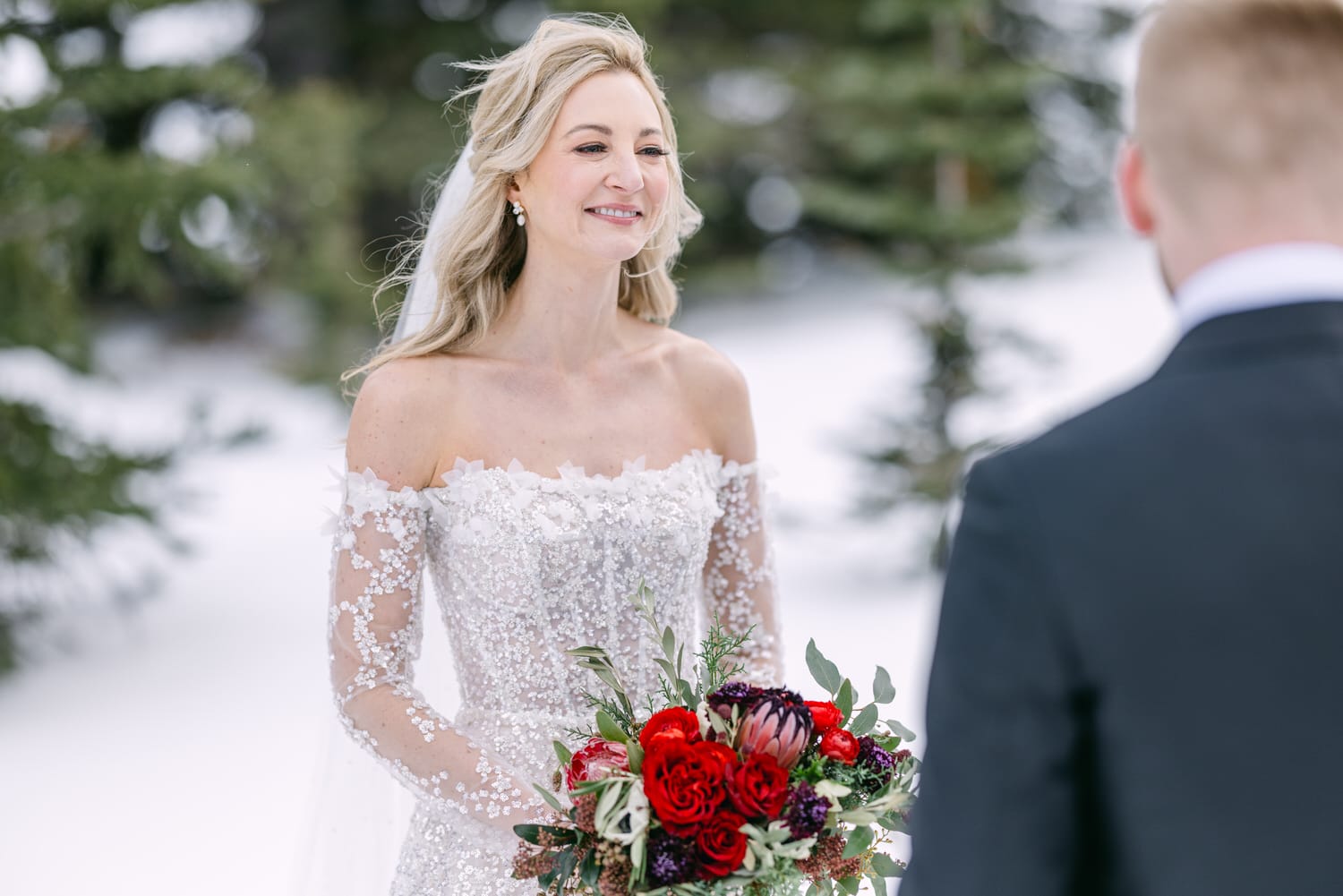 A bride in a white, sequined wedding dress holds a bouquet, smiling at the groom in a snowy forest setting.