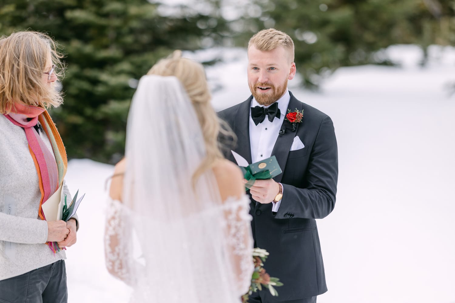 A groom reading his vows to a bride during a winter wedding ceremony with an officiant in the foreground.