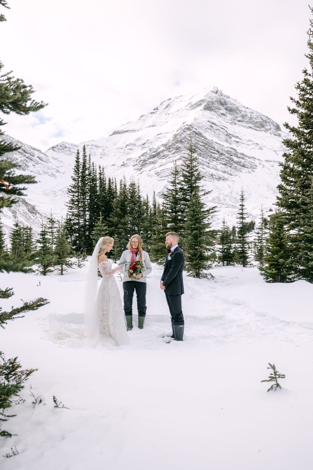 A bride and groom exchanging vows outdoors with an officiant in a snowy mountain setting.