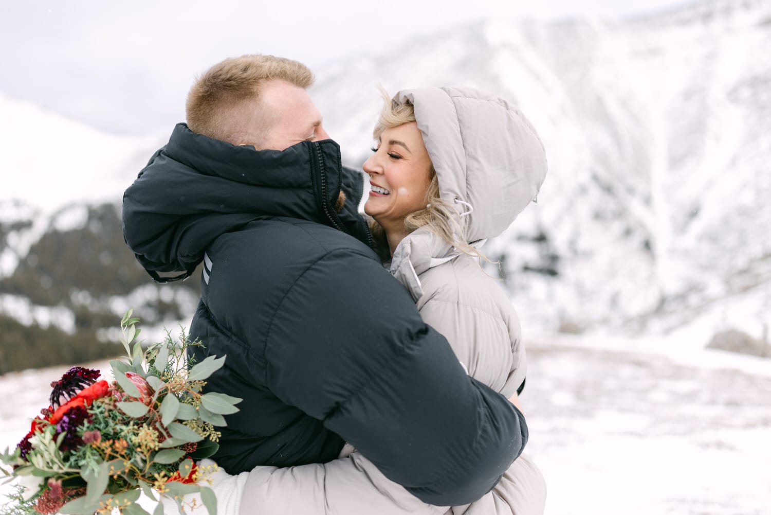 A man and woman happily hugging each other in a snowy landscape with the woman holding a bouquet of flowers.