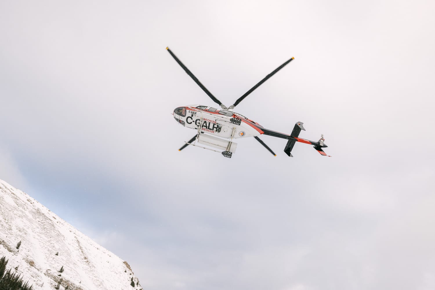 A red and white search and rescue helicopter flying with rotating blades against a cloudy sky, above a snow-covered mountain slope.