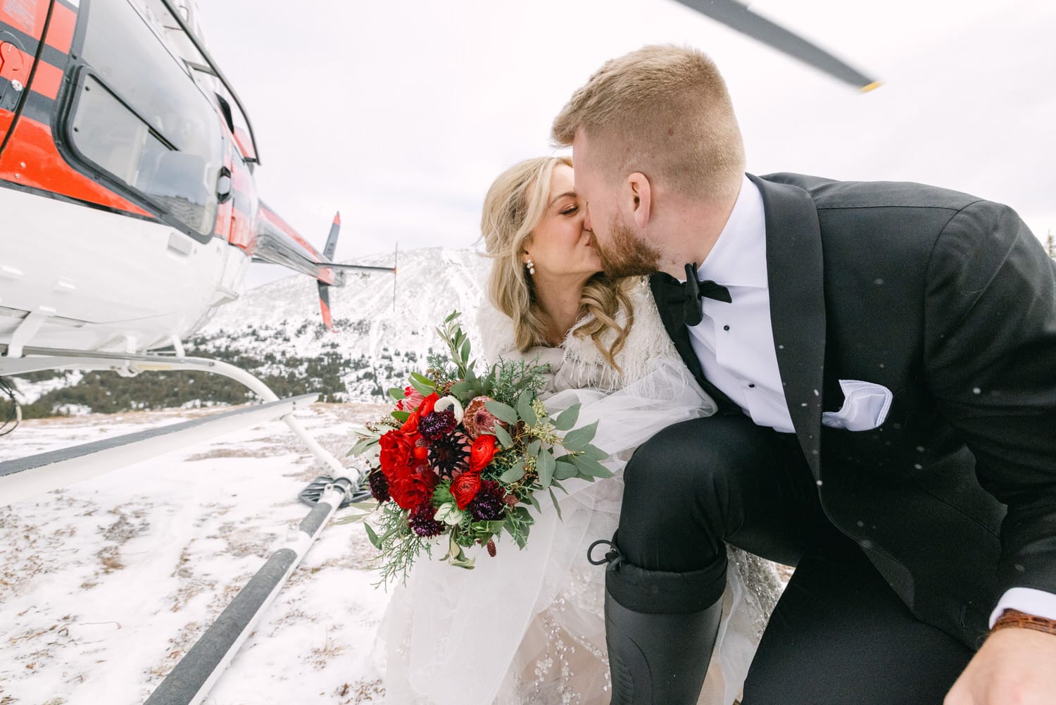 A bride and groom sharing a kiss in front of a red and white helicopter on a snowy mountain landing pad.