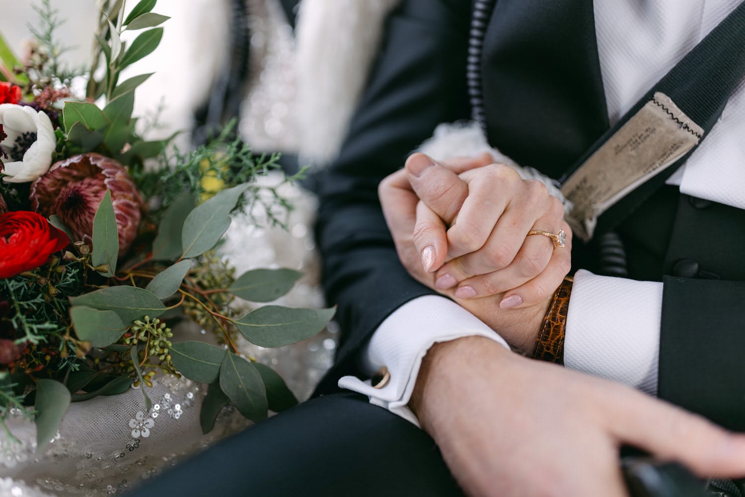 Close-up of a bride and groom holding hands during a wedding ceremony, with focus on the bride's ring and the couple's clasped hands, against a backdrop of a bridal bouquet and groom's suit.