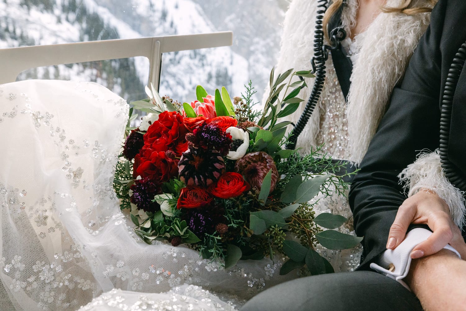 A bride in a sequined wedding dress with a fur shawl holds a bouquet of red flowers with snowy mountains in the background.