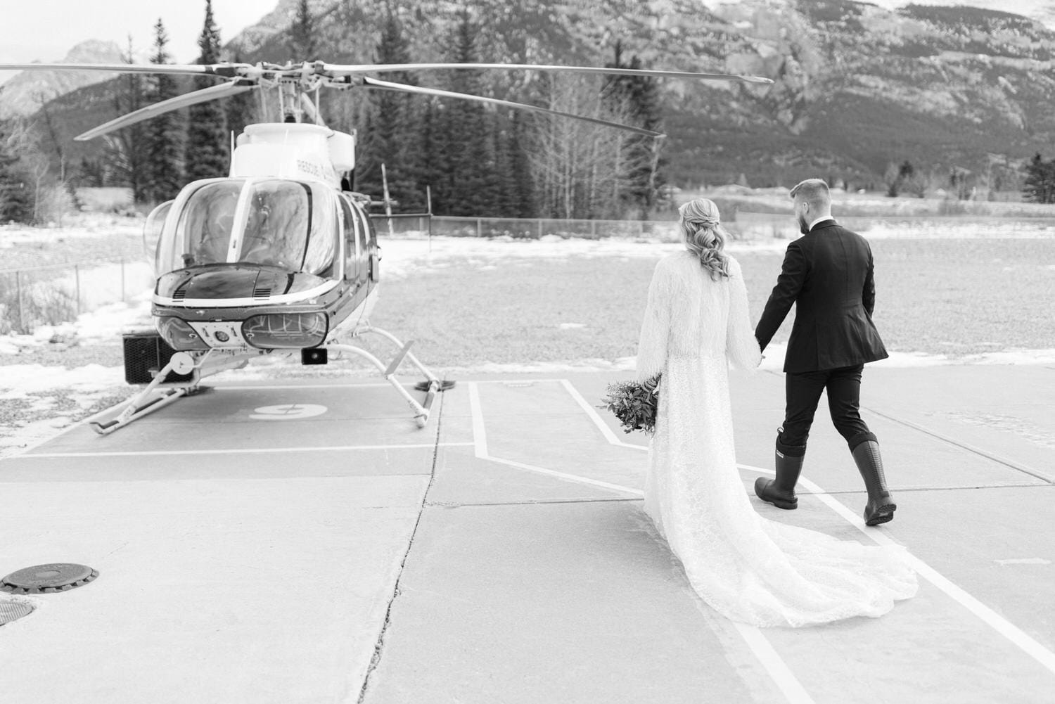 Black and white photo of a bride and groom holding hands and walking towards a helicopter, with mountains in the background.