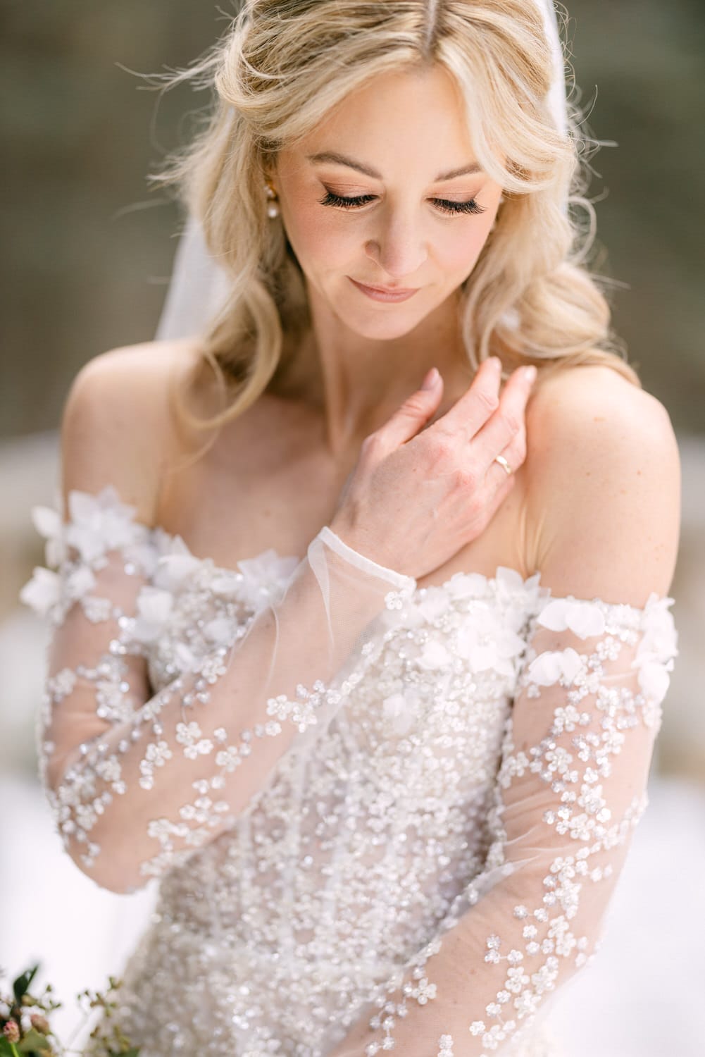 A bride in a white wedding dress with delicate floral embellishments, looking down and touching her shoulder gently.