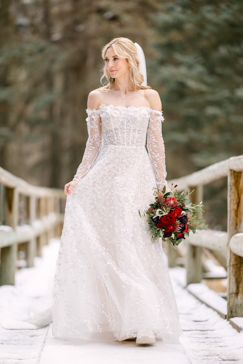 A bride in a long-sleeved, off-the-shoulder wedding gown carrying a bouquet of red flowers stands on a snowy wooden footbridge, surrounded by winter trees.