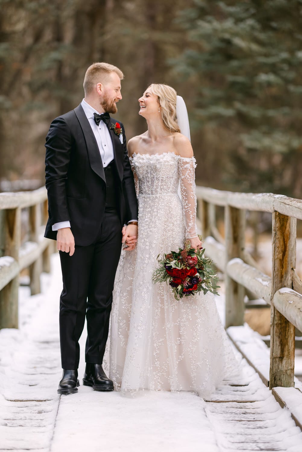 A bride and groom smiling at each other, dressed in elegant wedding attire, standing on a snowy wooden bridge with trees in the background.