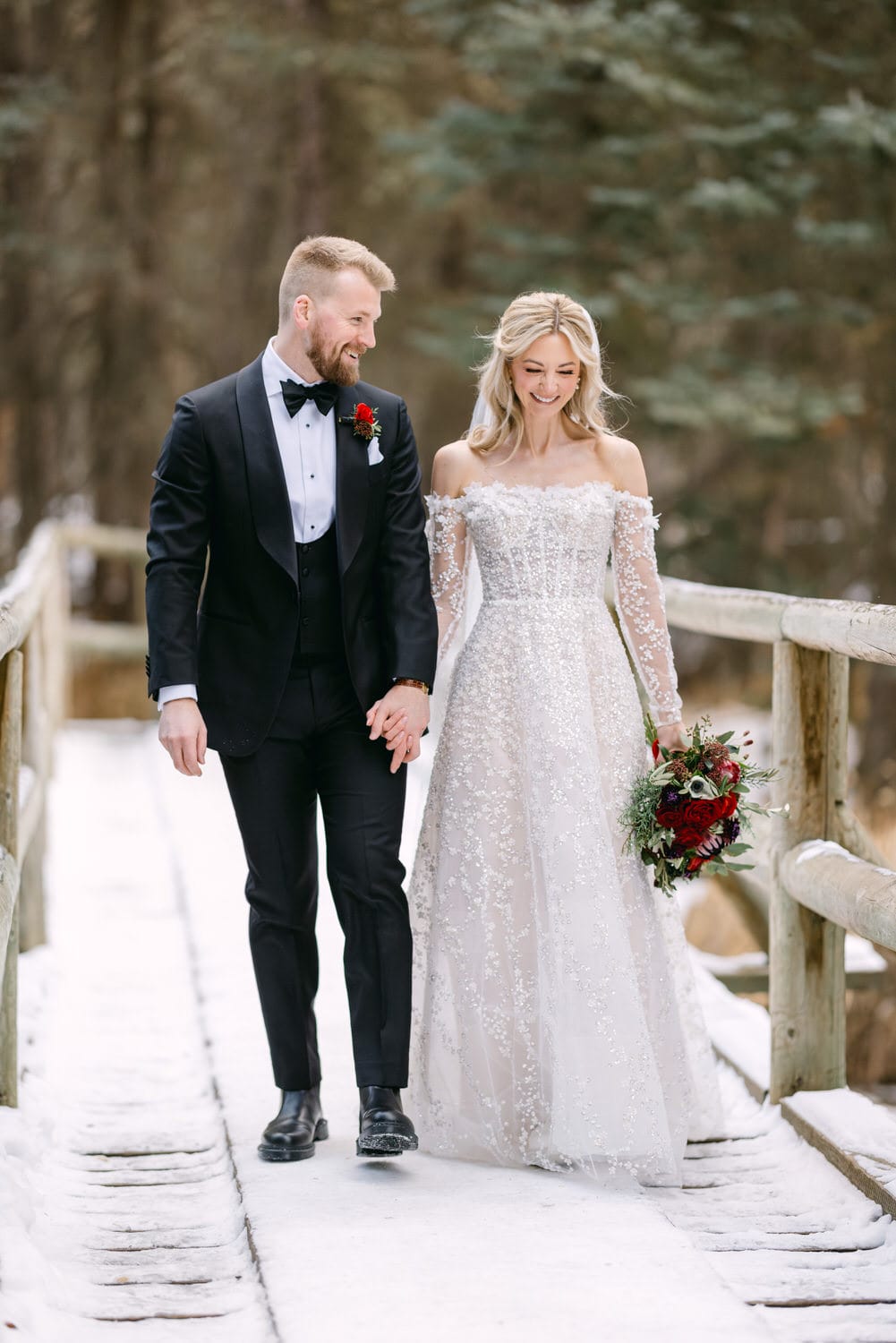 A bride and groom walking hand in hand on a snowy wooden bridge, surrounded by a forest.