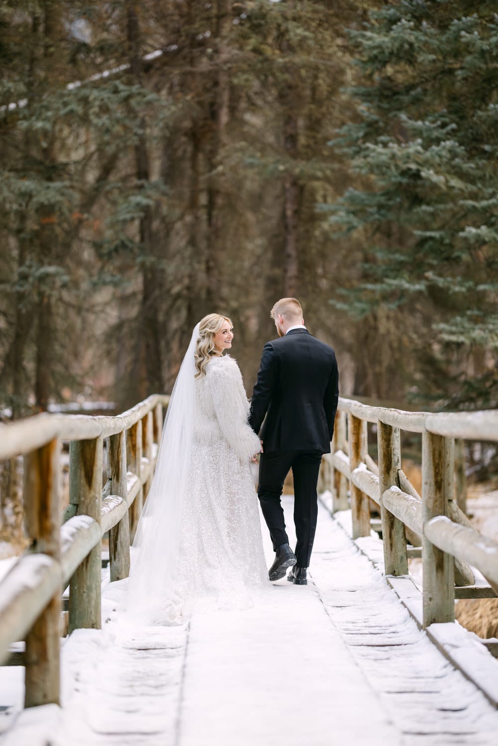 A bride in a white lace dress and a groom in a black suit walking hand in hand on a snowy wooden bridge surrounded by evergreen trees.