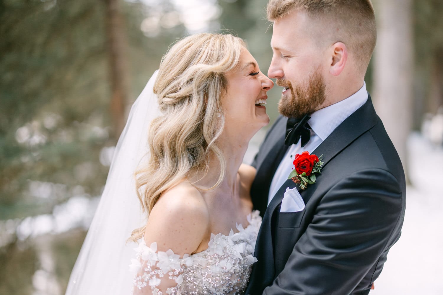 A bride and groom smiling and embracing each other in a forest setting, both dressed in wedding attire with a focus on their joyful expressions.