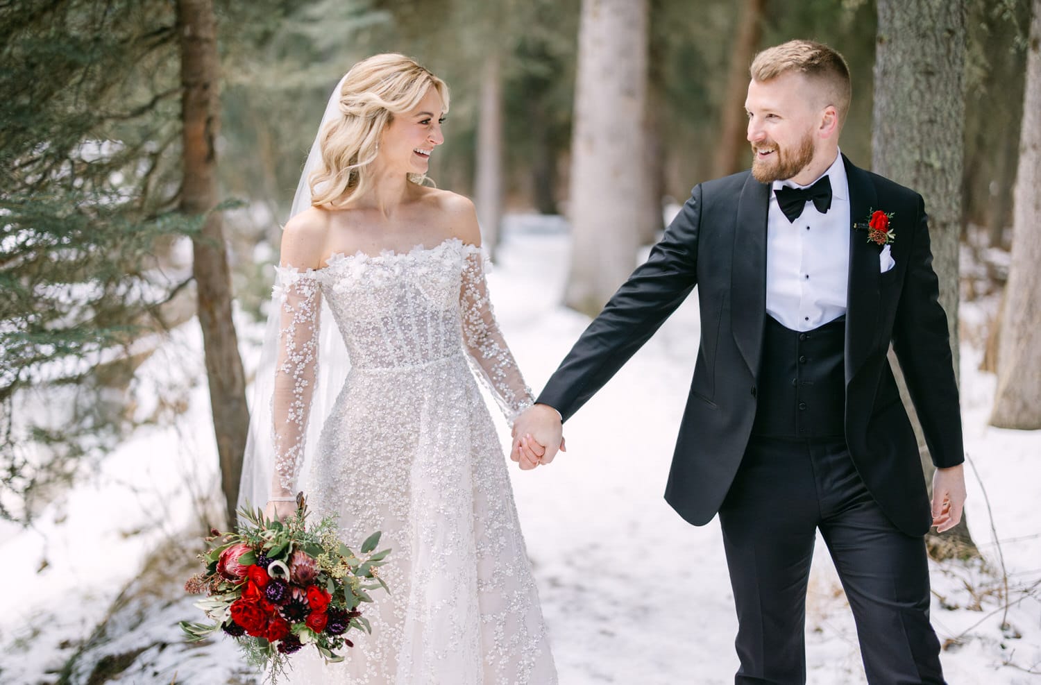 A bride and groom holding hands and smiling at each other in a snowy forest setting, with the bride carrying a bouquet of red flowers.