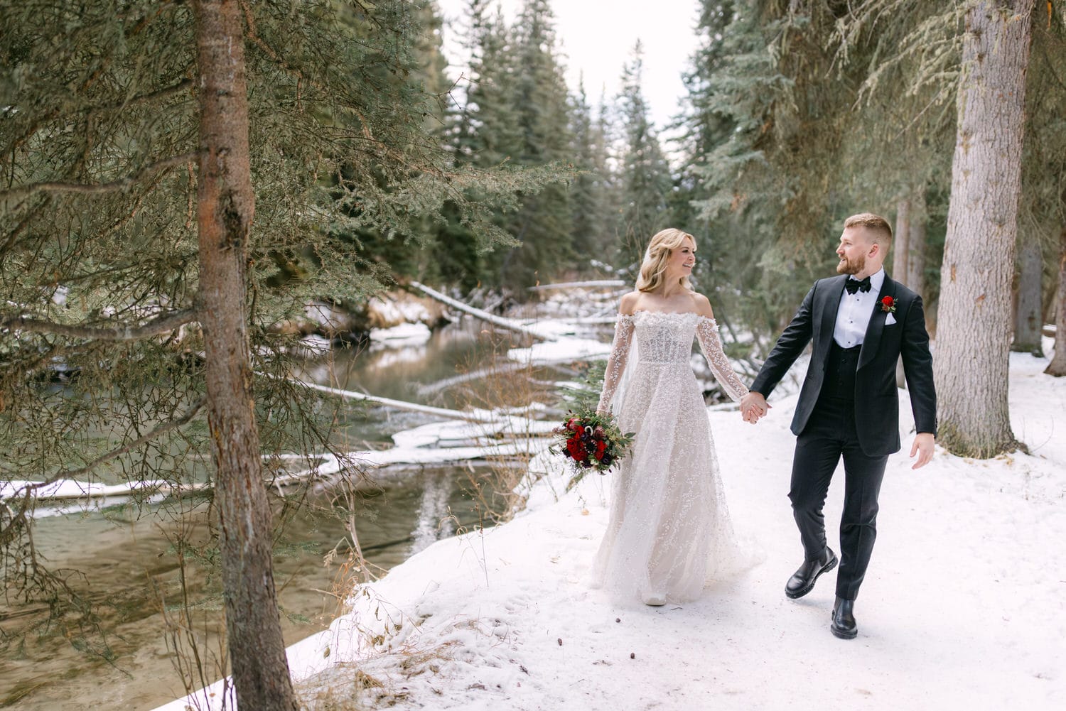 A bride and groom holding hands and walking through a snowy forest, with the bride carrying a bouquet and both dressed in wedding attire.