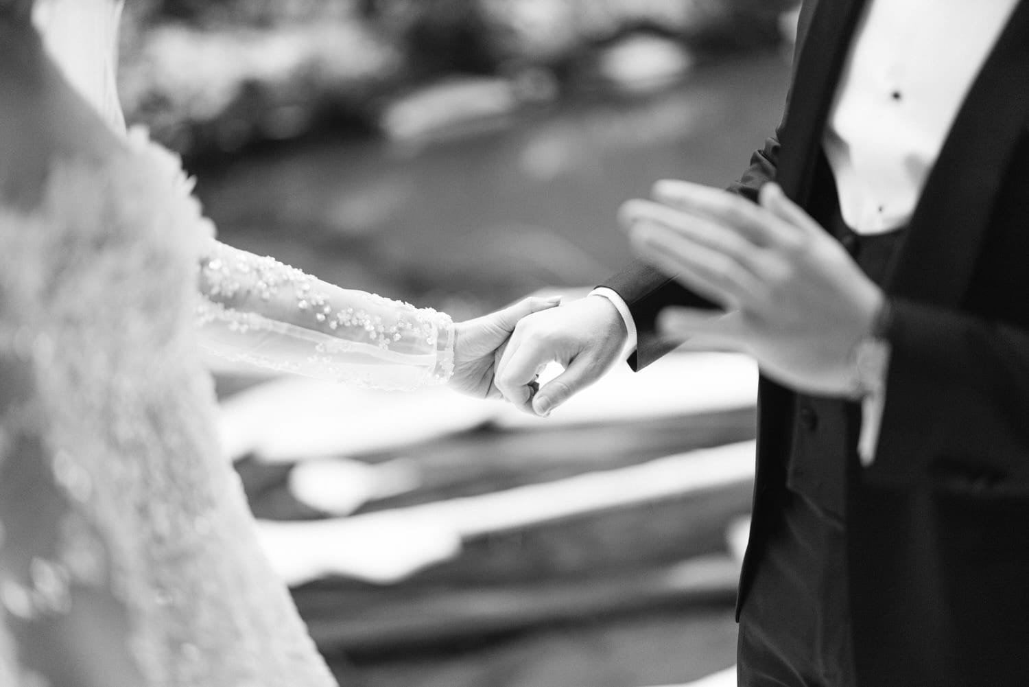 A black and white photo capturing a bride and groom holding hands on their wedding day, focusing on their hands with blurred background.