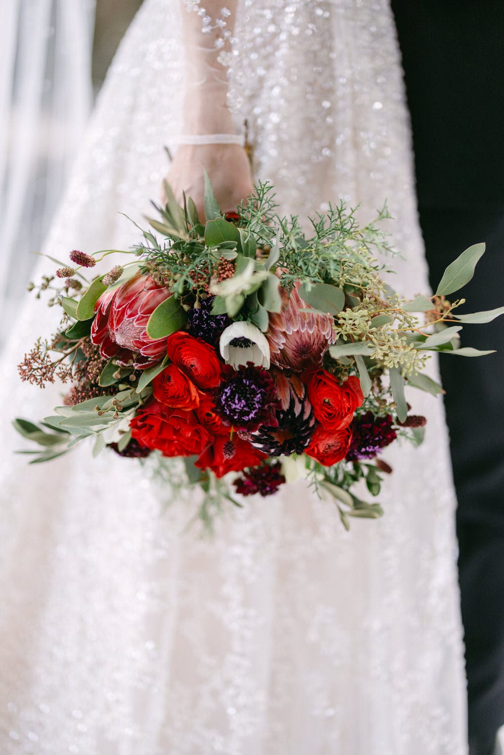 A bride holding a beautiful bouquet featuring red flowers and greenery, with a sparkling white wedding dress in the background.