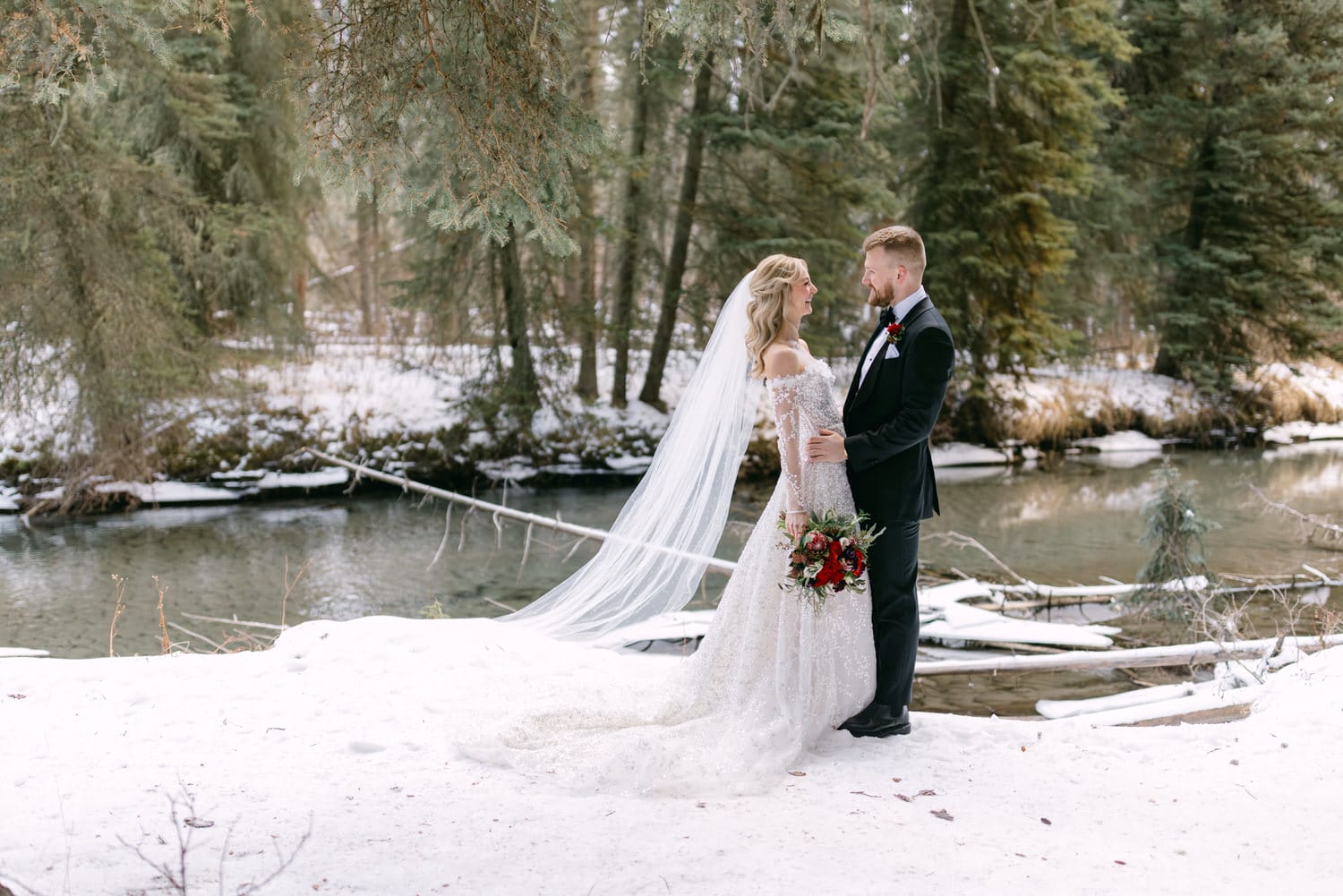 Bride and groom holding hands in a snowy forest setting with a stream in the background