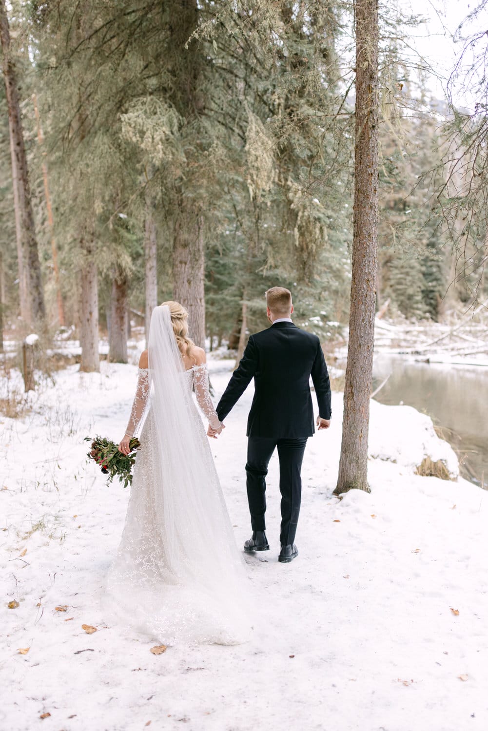 A bride and groom walking hand in hand through a snowy forest setting.