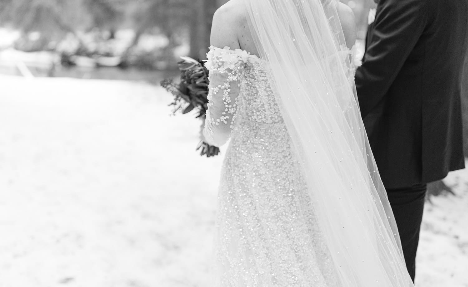 Close-up of a bride in a detailed wedding dress and a groom, in black and white, with the focus on the bride's bouquet and the sparkling details of her dress.