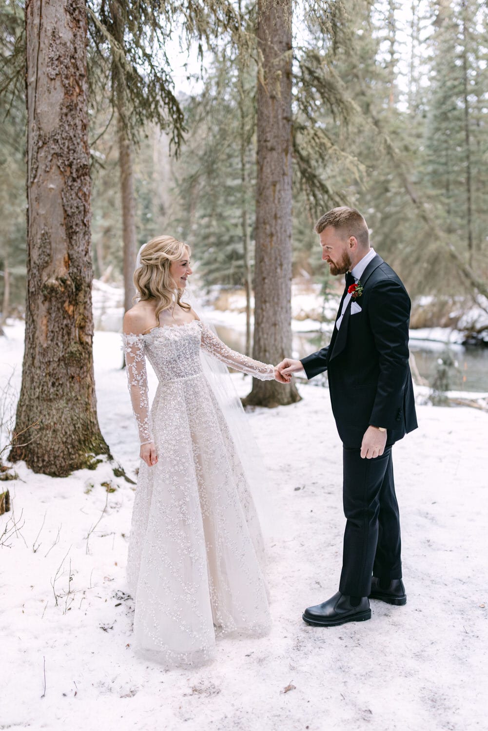 Bride in a lace wedding dress holding hands with groom in a black suit in a snowy forest setting