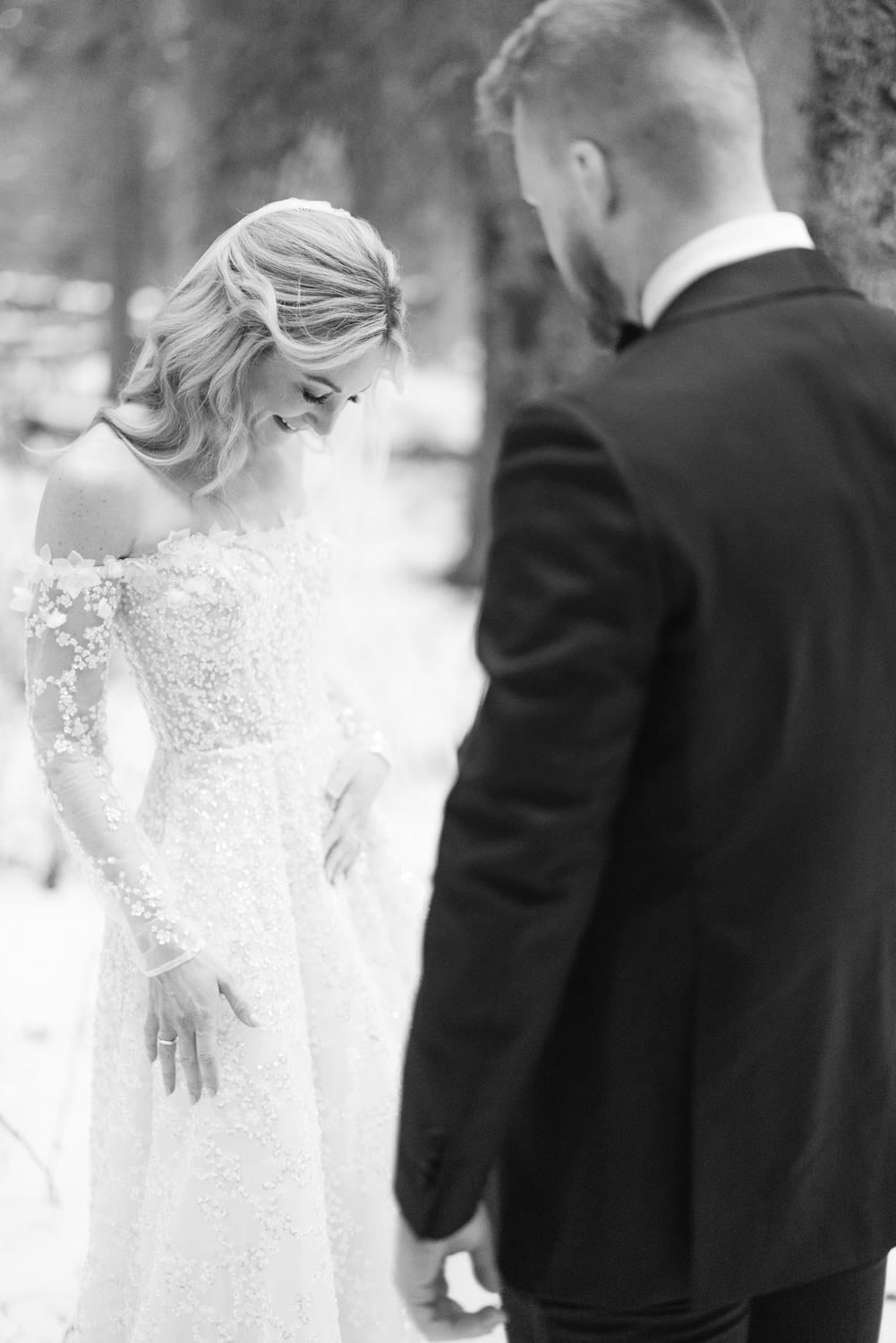 Black and white photo of a bride in a lace gown smiling downwards, with a groom standing in profile to her, both in a forest setting.