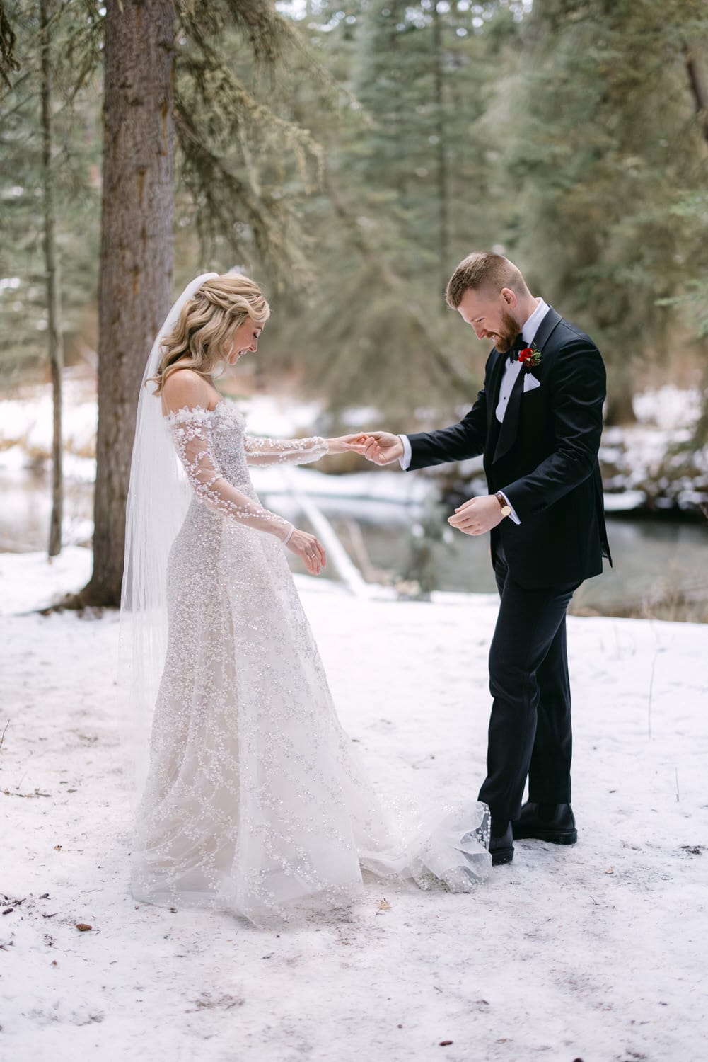 Winter Wonderland Wedding:::A bride in a white laced gown and a groom in a black suit holding hands in a snowy forest setting.