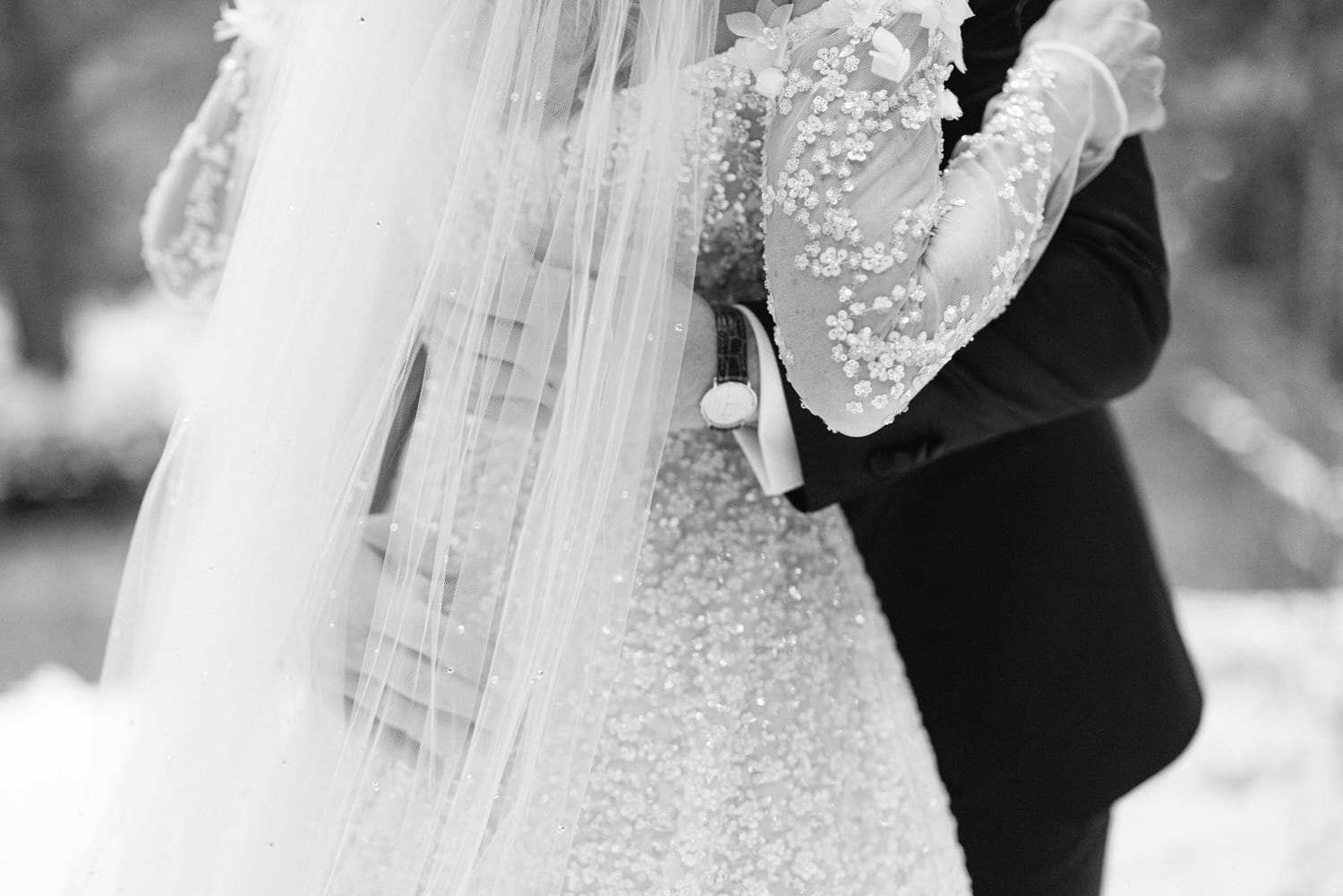 Close-up black and white photo of a bride and groom embracing, focusing on the bride's beaded dress and veil.