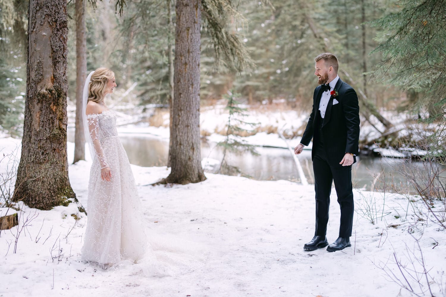 A bride in a lace dress and a groom in a black tuxedo standing in a snowy forest setting.